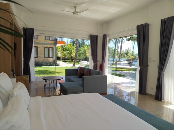 A hotel room with a large bed, a sofa, a small table, and a view of a pool and palm trees. - Scampi Indorus Resort (Tejakula)