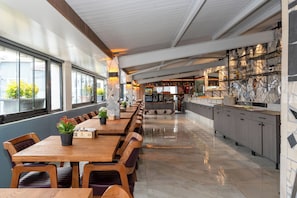 Un restaurant moderne doté d’une longue table à manger en bois, de chaises rembourrées et d’un espace cuisine avec des plans de travail en marbre.