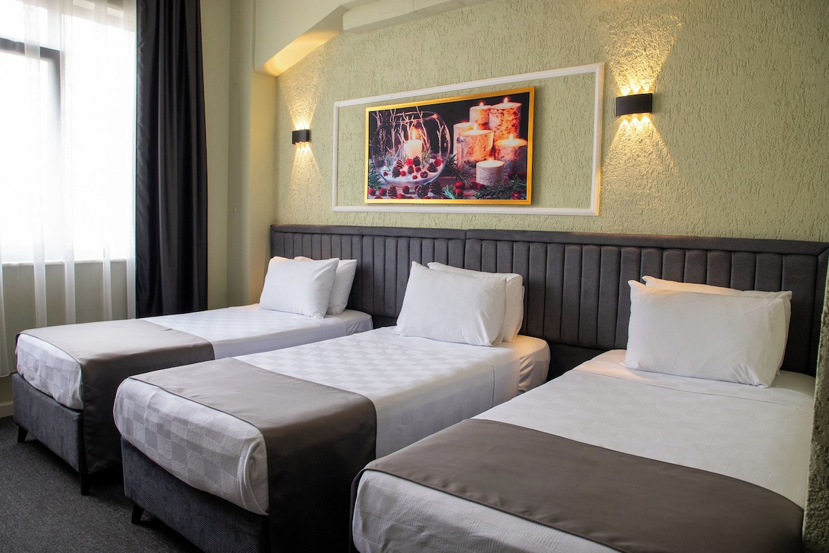 Une chambre d’hôtel avec trois lits simples, un tableau au mur et une fenêtre avec des rideaux.