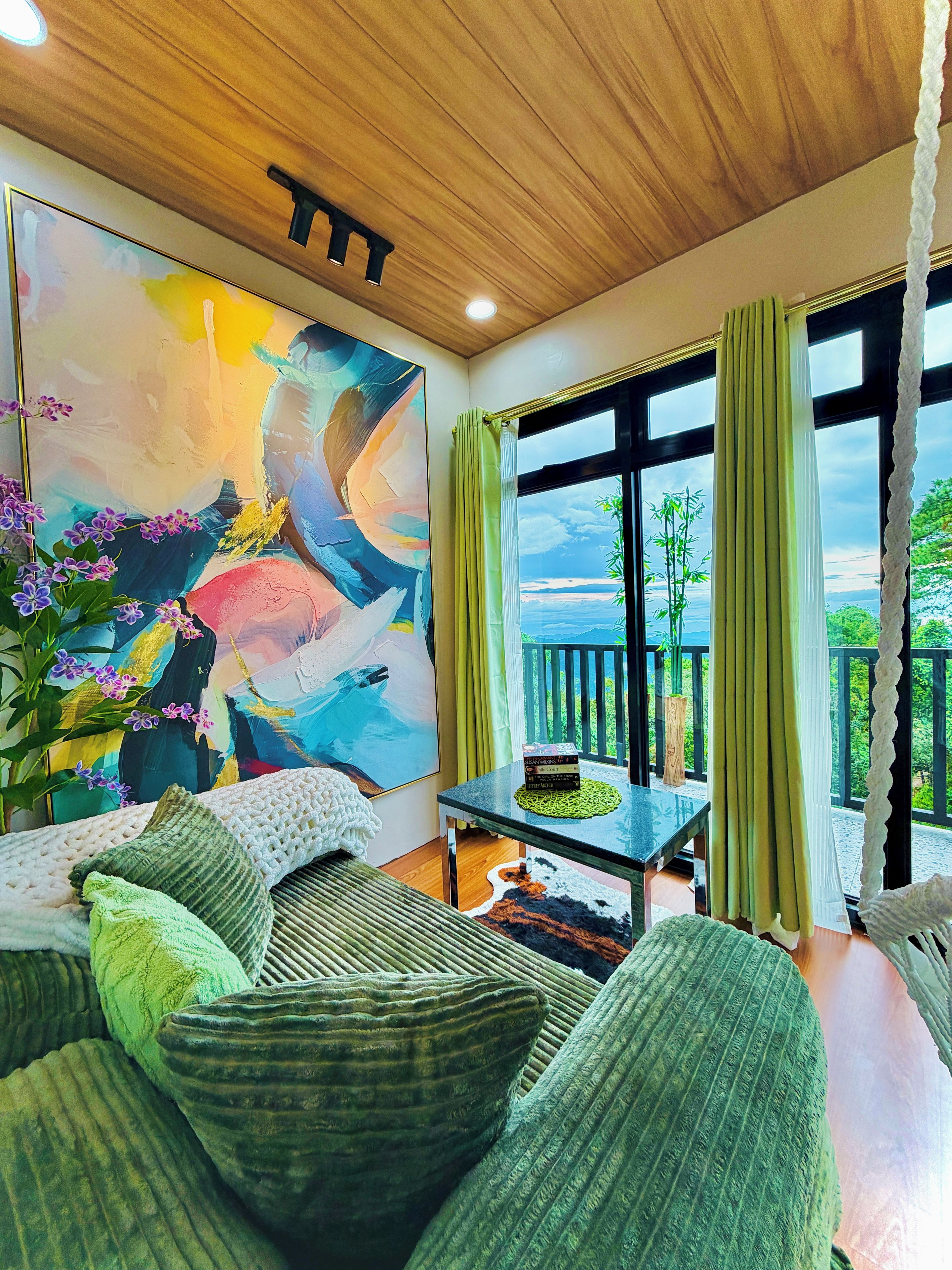 A room with a large abstract painting, a green sofa, a coffee table, and a view of greenery outside.