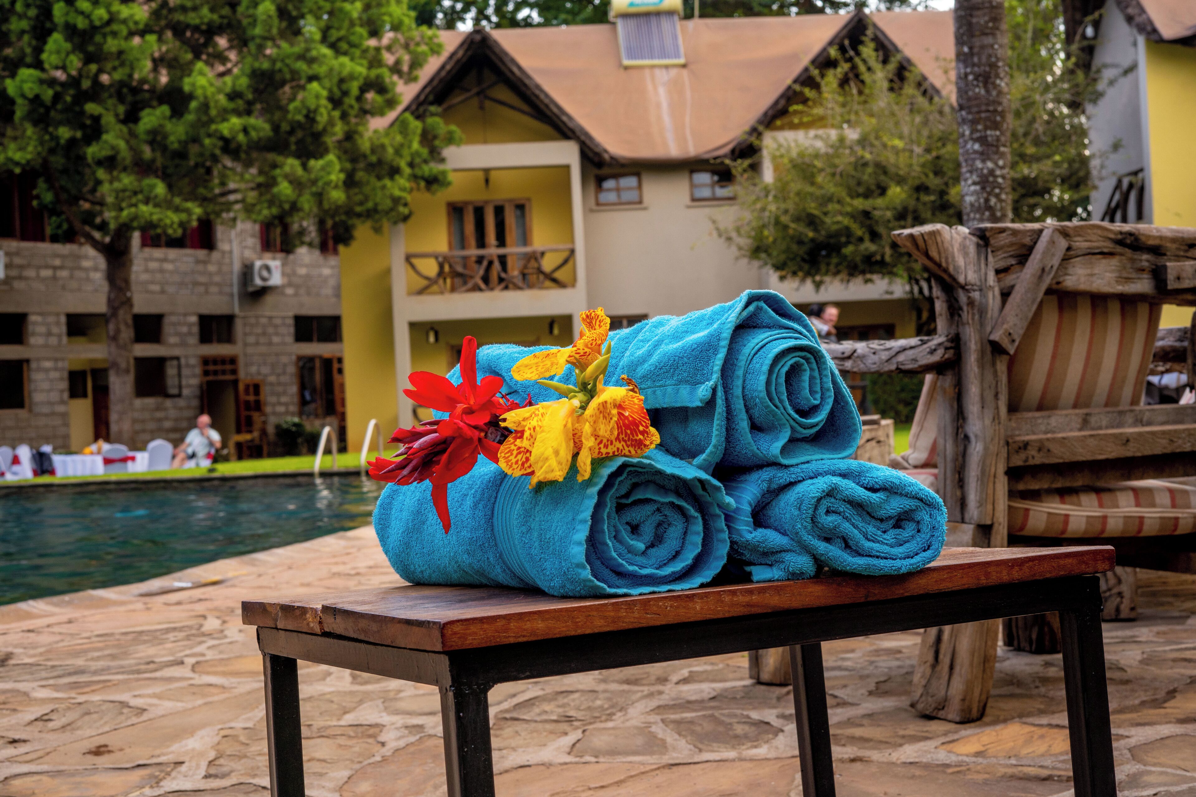 A poolside area with a wooden table holding rolled towels and a bouquet of flowers.