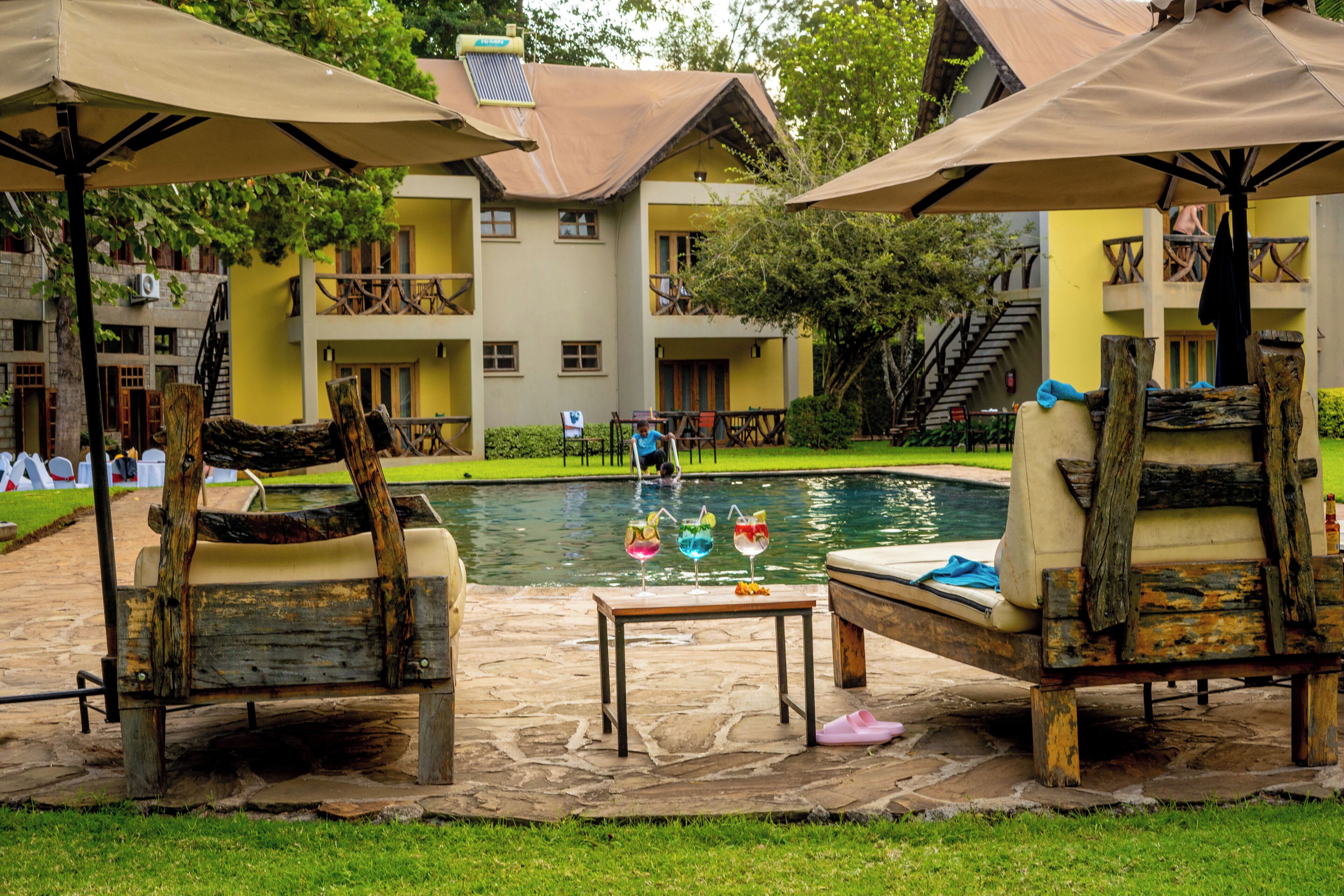 A poolside area with lounge chairs and umbrellas, a stone table with drinks, and a building with balconies in the background.
