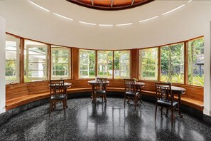 Una habitación con un banco de madera curvo, cuatro mesas redondas y ventanas amplias con vistas a zonas verdes.