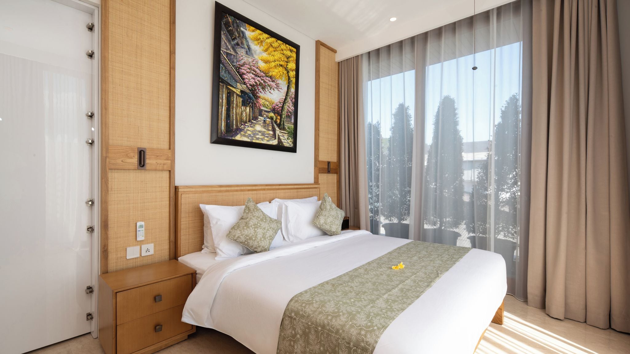 A hotel room with a large bed, a wooden nightstand, a painting on the wall, and a view of trees through the window.