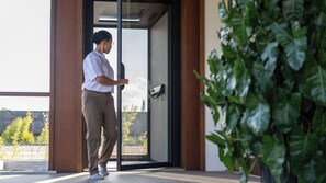 A person standing at a glass door with a handle, a plant to the right, and a view of trees and a building outside. - The Atma Bali (Denpasar)
