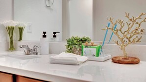 Casa de banho com lavatório branco, espelho, suporte de papel higiénico, planta verde e uma árvore decorativa.