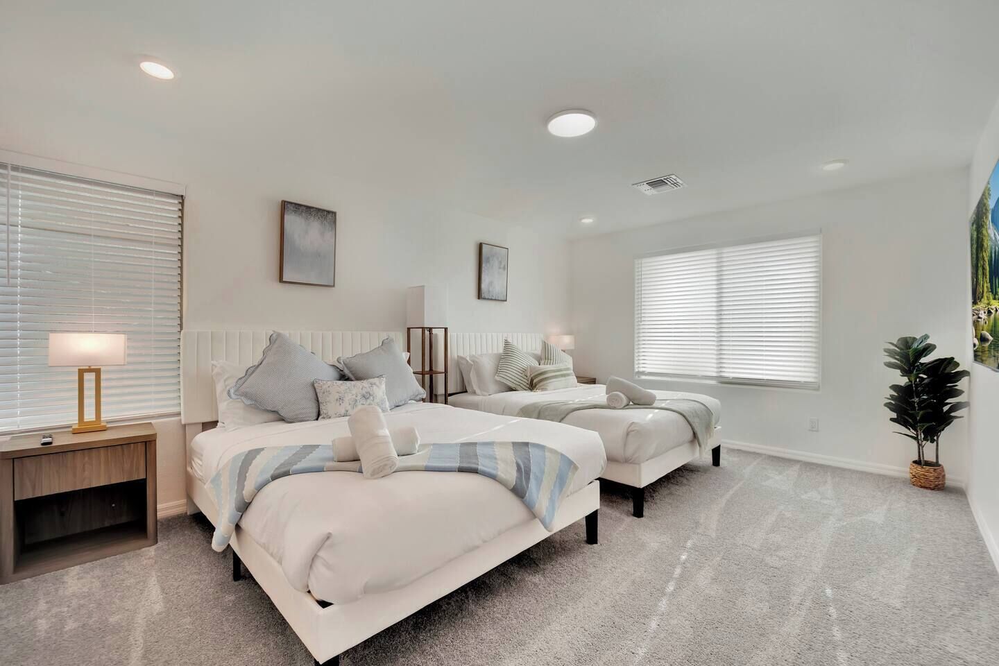 A bedroom with two beds, a bedside table with a lamp, a radiator, and a large window with blinds.