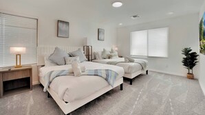 Quarto com duas camas, mesa de cabeceira com luminária, radiador e uma janela grande com persianas.