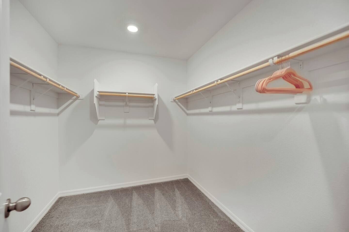 Habitación pequeña con paredes blancas, techo inclinado y suelo alfombrado. Se observan dos estantes de madera con ropa colgada y un solo perchero rosa.