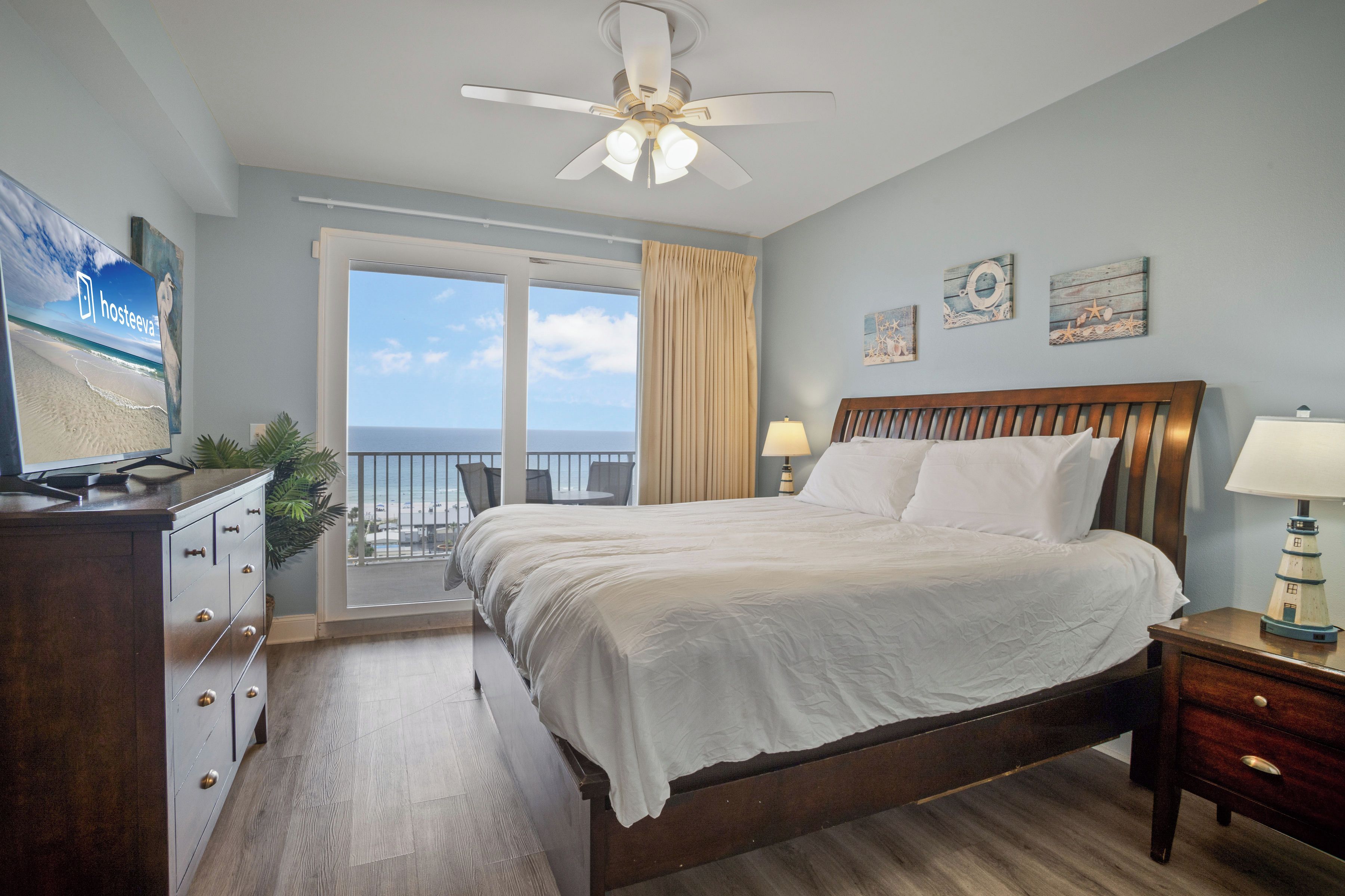 A hotel room with a bed, a desk, a chair, a nightstand, a lamp, a ceiling fan, and a view of the ocean through a large window.