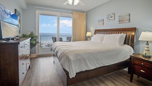A hotel room with a bed, a desk, a chair, a nightstand, a lamp, a ceiling fan, and a view of the ocean through a large window. - Laketown Wharf 824 (Panama City Beach)