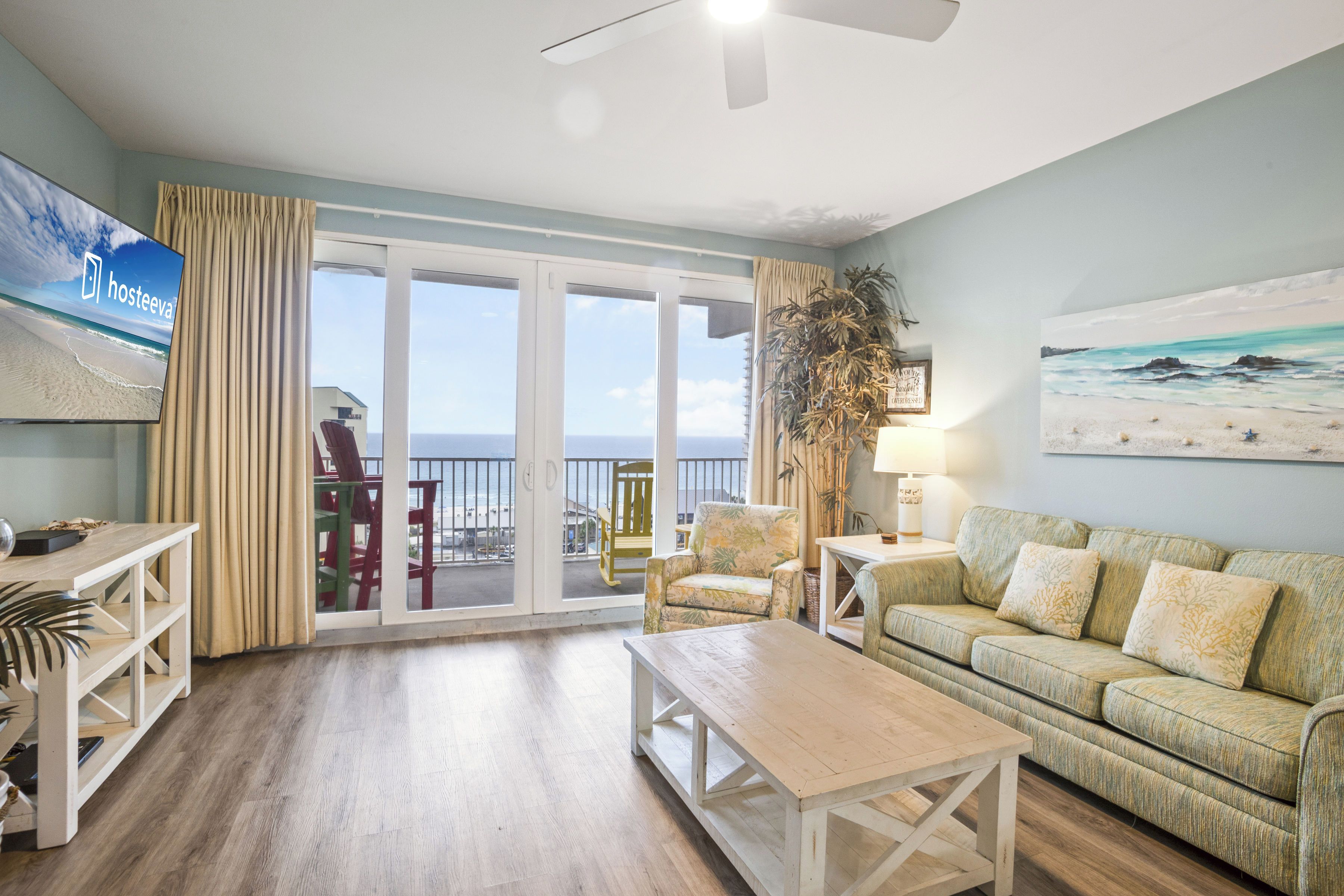 A living room with a wooden floor, a sofa, a coffee table, a TV, and a balcony with a view of the beach.