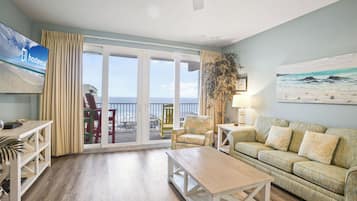 A living room with a wooden floor, a sofa, a coffee table, a TV, and a balcony with a view of the beach.
