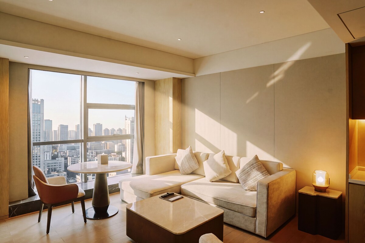 Quarto de hotel moderno com sofá, mesa de centro redonda e vista para a paisagem urbana através de grandes janelas.