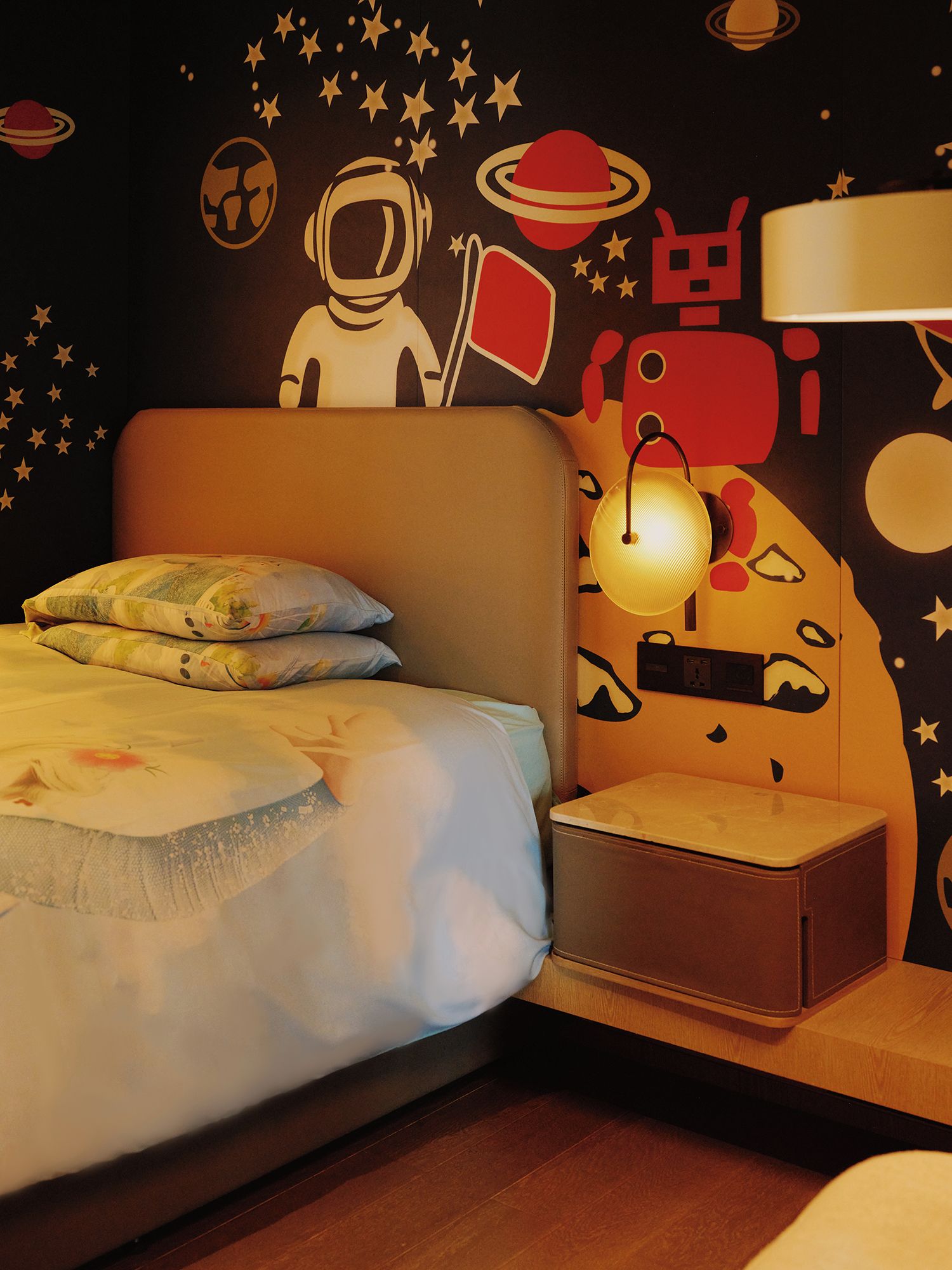 A bedroom with a bed, pillows, a nightstand, a lamp, and a wall with space-themed decorations.