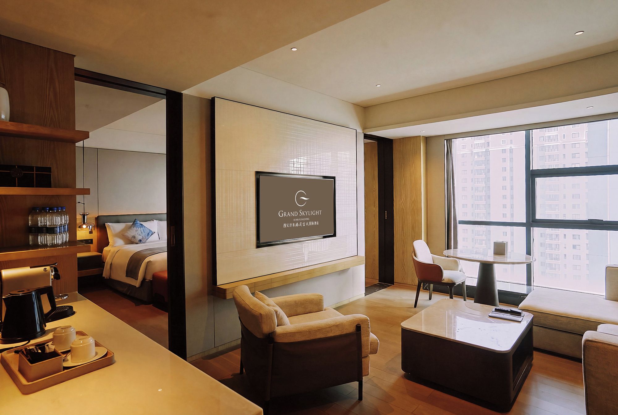 A hotel room with a large window, a flat-screen TV mounted on the wall, a sofa, a coffee table, and a desk with a coffee machine.