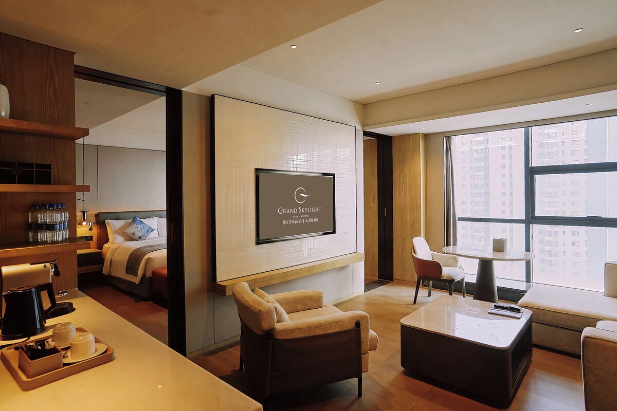 Habitación de hotel con un ventanal grande, un televisor de pantalla plana en la pared, un sofá, una mesa de centro y un escritorio con una cafetera.