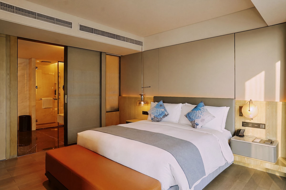 Habitación de hotel moderna con una cama grande, una mesita de noche con lámpara y un baño visible a través de una puerta corredera.