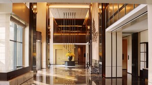 Un moderno vestíbulo de hotel con piso reflectante, una columna central con un jarrón de flores y una escalera a la derecha.