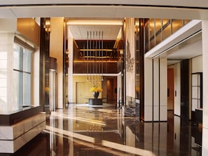 Um hall de hotel moderno com piso refletivo, uma coluna central com um vaso de flores e uma escadaria à direita.