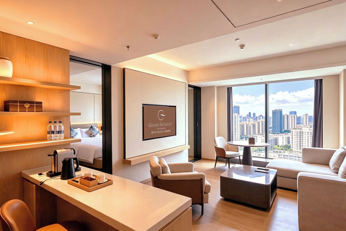 Quarto de hotel moderno com cama, escrivaninha com cafeteira, sofá, poltrona e grande janela com vista para a cidade.
