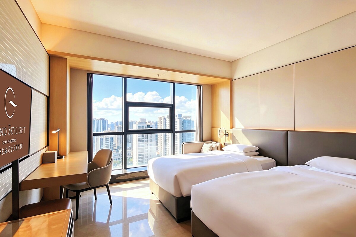 Quarto de hotel com duas camas, uma escrivaninha, uma cadeira e uma janela grande com vista para a cidade.