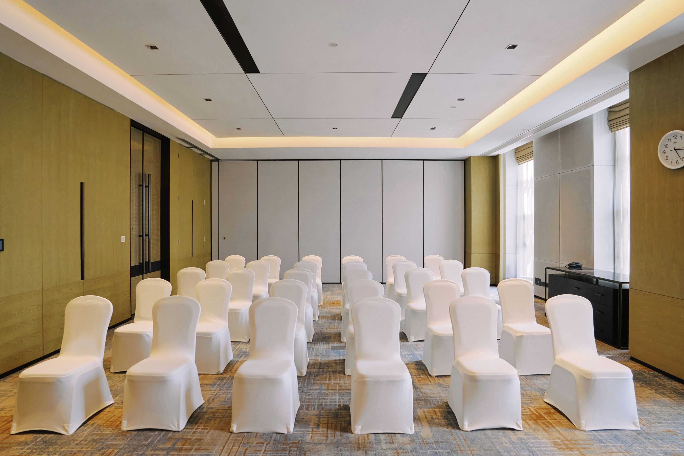 A conference room with rows of white chairs, a clock on the wall, and a carpeted floor.