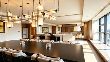 Habitación de hotel moderna con una amplia mesa de comedor, varias opciones de asiento, una zona de cocina y vistas a la ciudad a través de grandes ventanales.