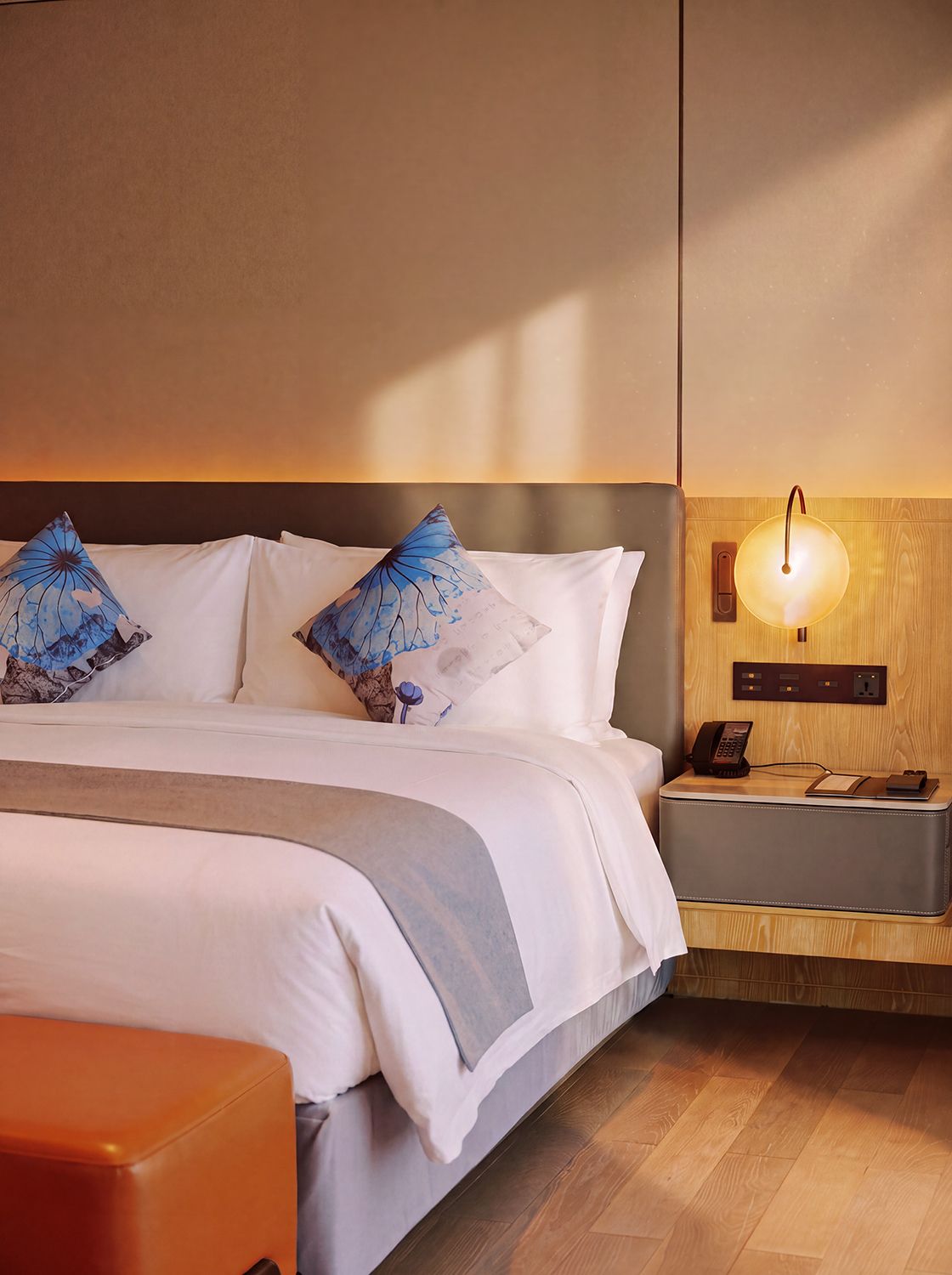 Habitación de hotel con cama, almohadas, mesita de noche, lámpara y suelo de madera.