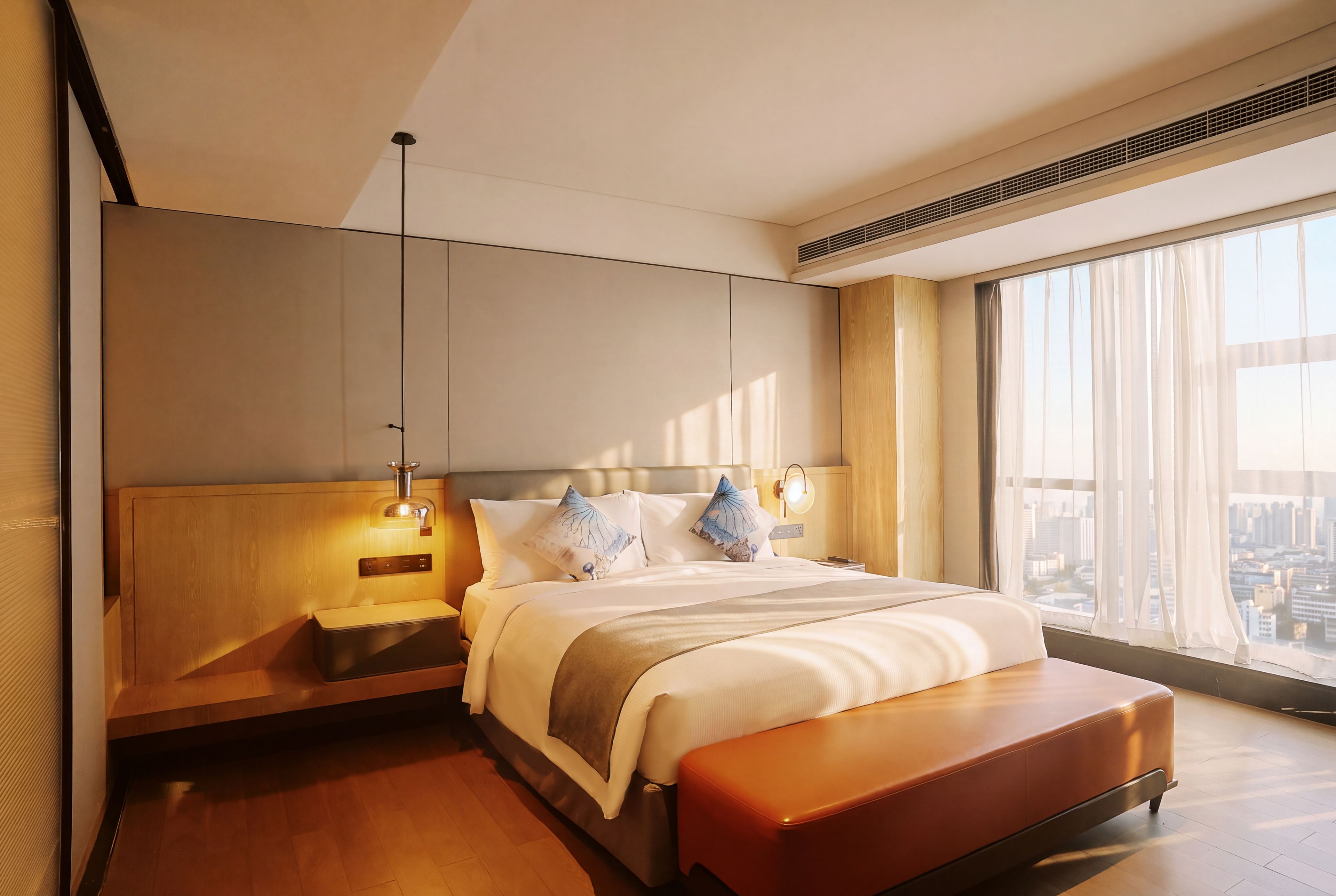 Una habitación de hotel moderna con una cama grande, un banco de madera, una mesita de noche y un ventanal con vistas a la ciudad.