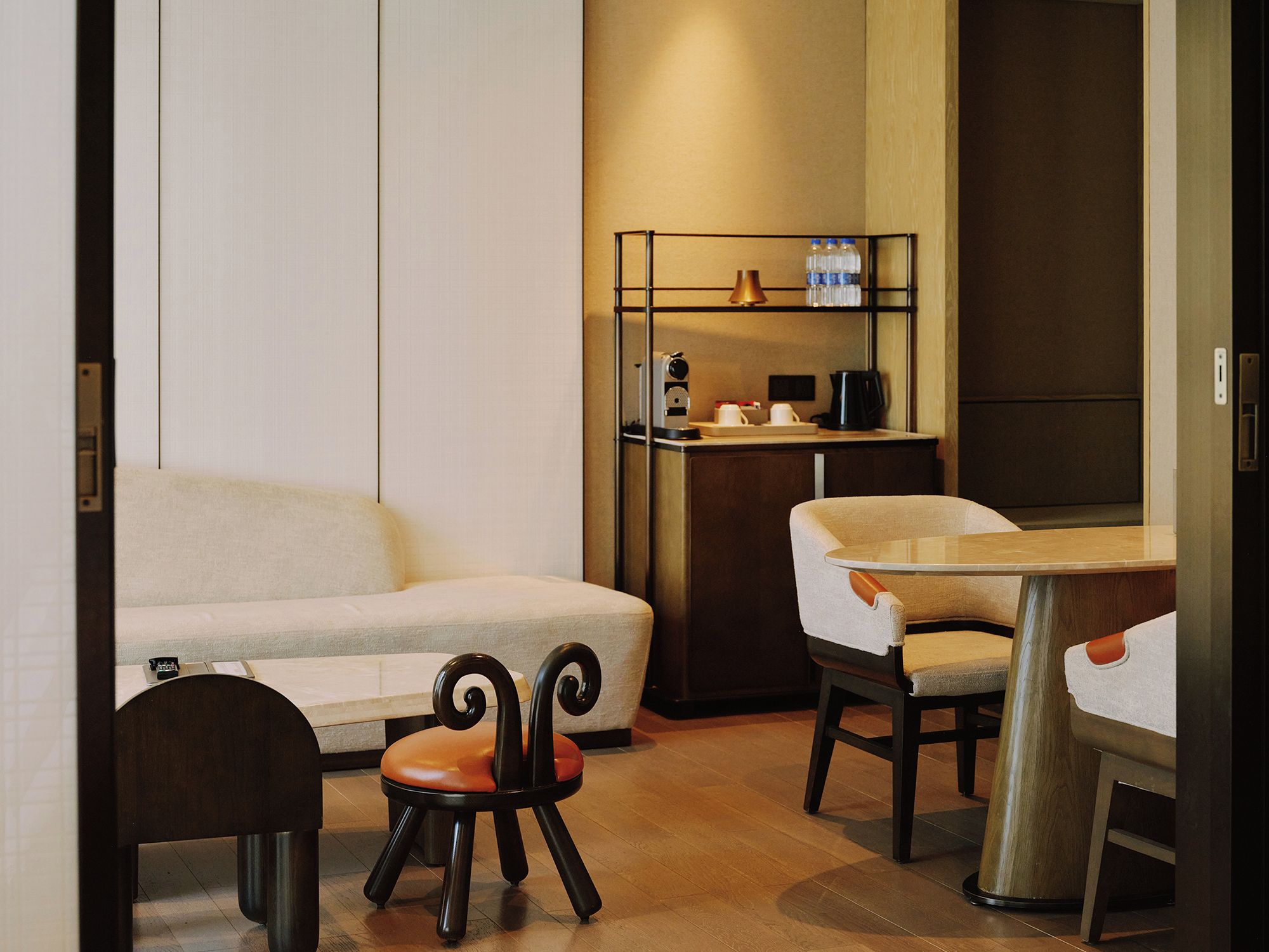 A modern hotel room with a sofa, a dining table, and a small stool.