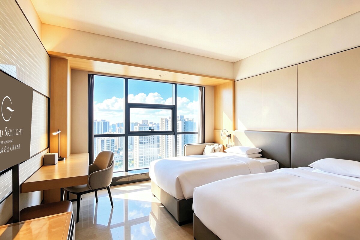 A hotel room with two beds, a desk, a chair, and a large window offering a city view.