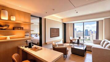 A modern hotel room with a bed, a desk with a coffee maker, a sofa, a chair, and a large window with a city view.