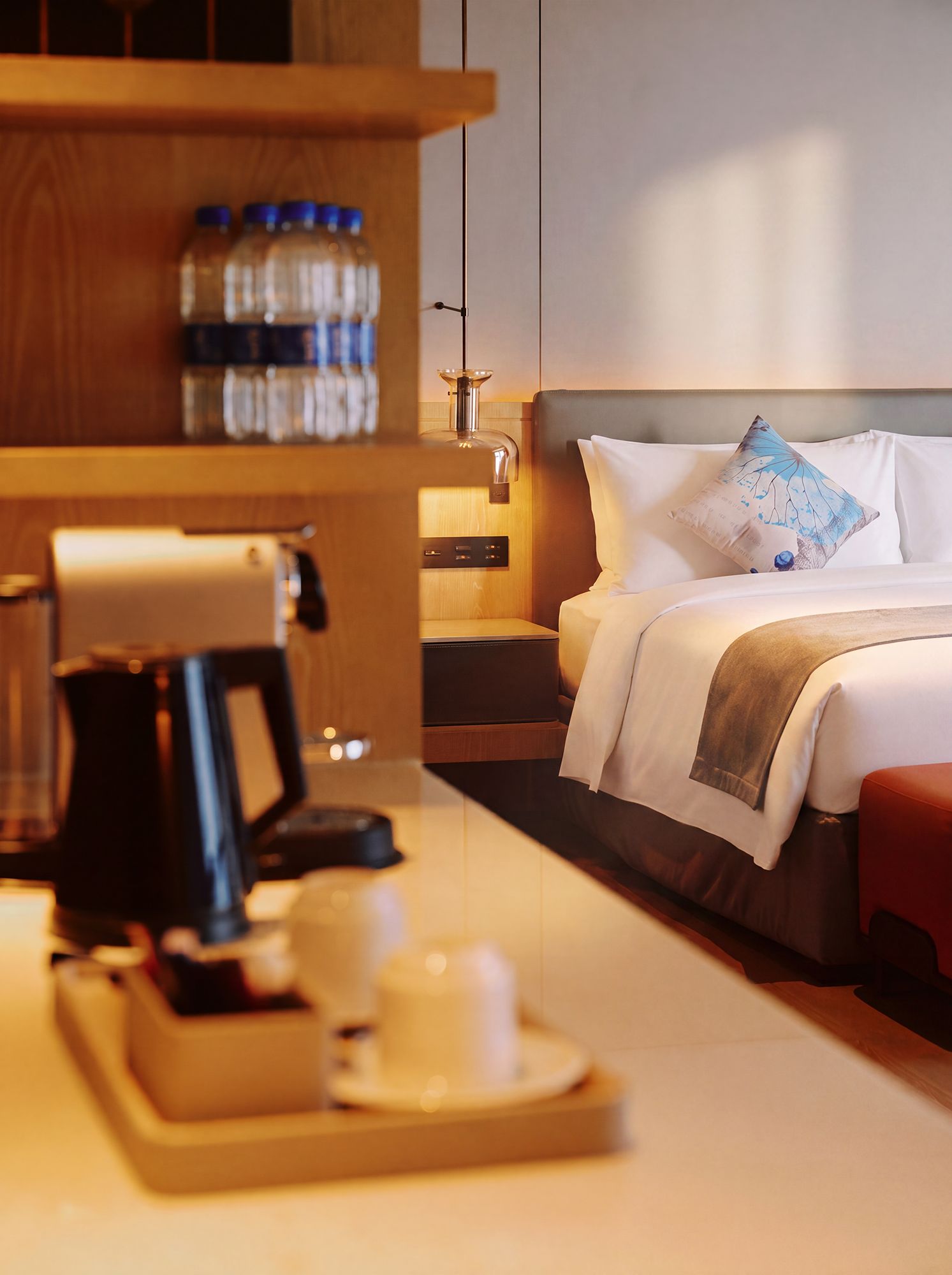 A hotel room with a bed, a bedside table with a coffee maker, a tray with a cup and saucer, and a shelf with bottles.