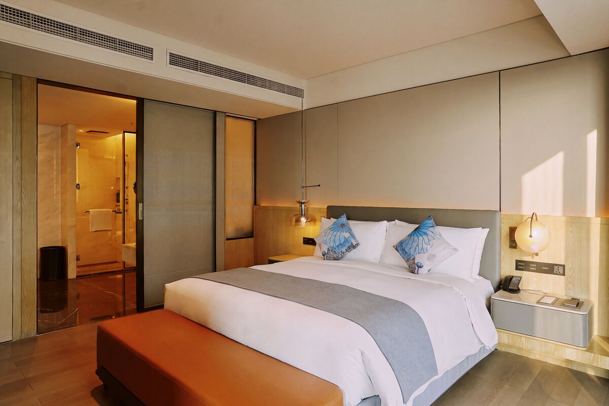 Quarto de hotel moderno com uma cama grande, mesa de cabeceira com abajur e banheiro visível por uma porta deslizante.