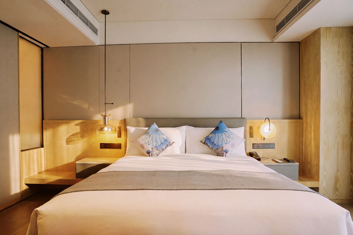 Quarto de hotel com uma cama grande, duas mesas de cabeceira, um criado-mudo, telefone fixado na parede e luminária no teto.