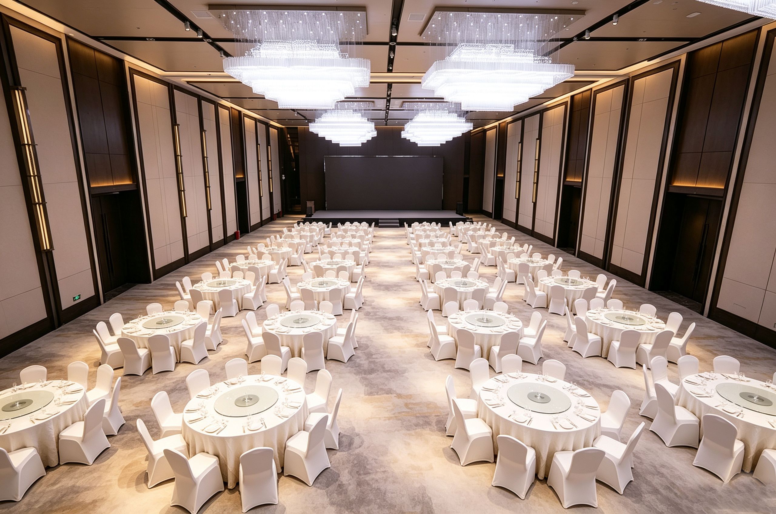 A long, narrow hotel room with rows of round tables and chairs, a stage at the far end, and modern chandeliers.