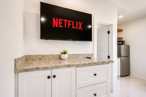Eine Küche mit einer Granitabdeckung, weißen Schränken, einem schwarzen Fernseher, der über der Arbeitsplatte 'NETFLIX' anzeigt, und einem Edelstahl-Kühlschrank.