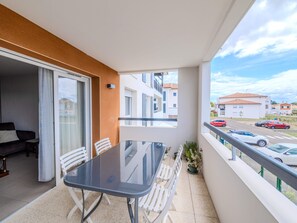 Un balcon avec une table et des chaises, une porte-fenêtre coulissante et une vue sur la rue où sont garées des voitures.