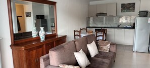 Una sala de estar con un sofá marrón, un aparador de madera y un espejo grande. Se ve una cocina con armarios blancos y un refrigerador al fondo.