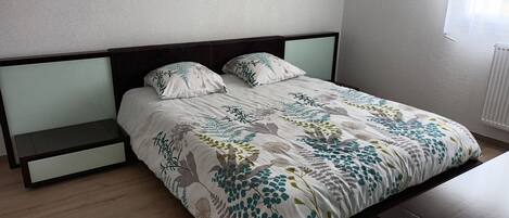 Una cama con una colcha estampada, dos almohadas, una mesita de noche y una mesita auxiliar.