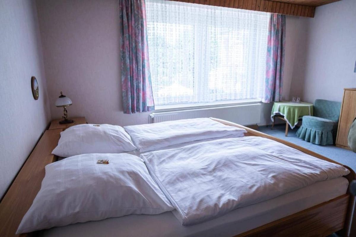 A double bed with white linens in a room with a window, a small table, and a chair.