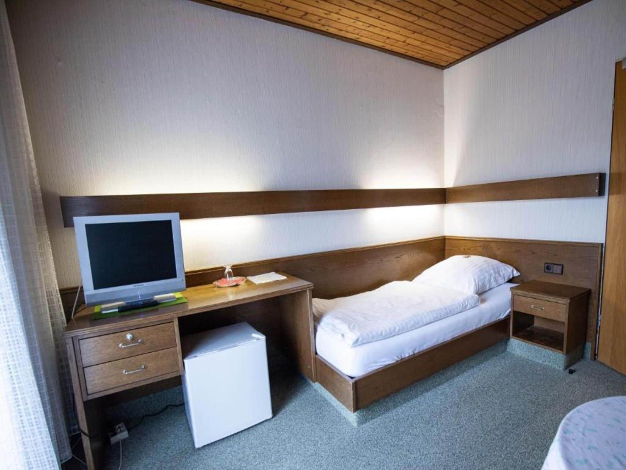 A compact hotel room with a single bed, a desk with a computer, a small refrigerator, and a bedside table.