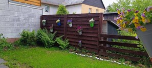 Una cerca de madera con macetas colgantes, un jardín césped y un edificio al fondo.
