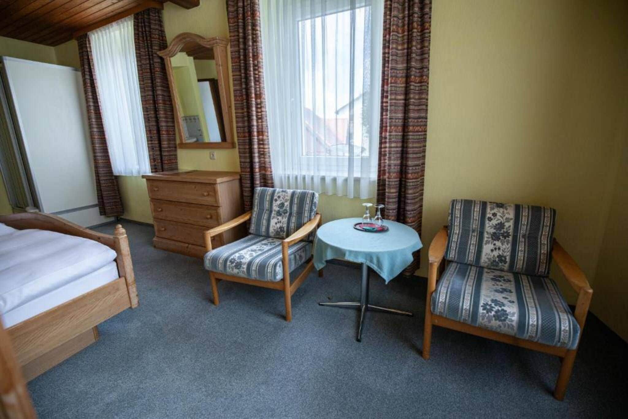 A room with a bed, two armchairs, a small table, a dresser, and a window with curtains.