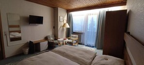 Una habitación de hotel con cama, escritorio, silla, lámpara y ventana con cortinas.