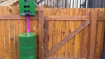 A dog waste station with a green trash can and a sign, a wooden fence, and a dog standing on a concrete slab.