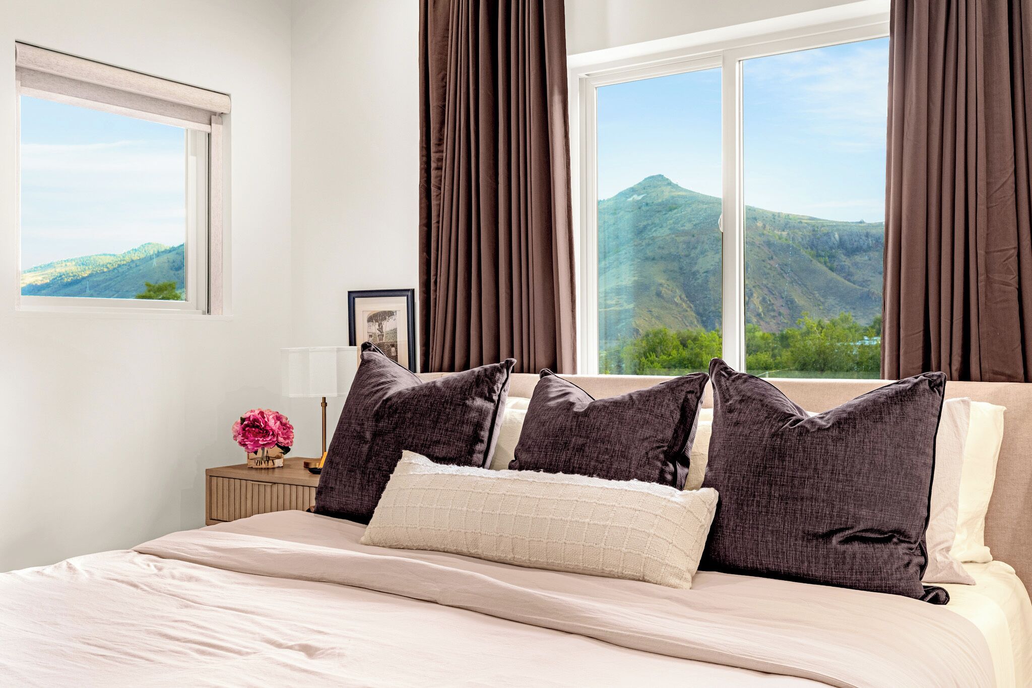 Une chambre à coucher avec un lit, une table de chevet, une lampe, un tableau encadré et une fenêtre avec des rideaux.