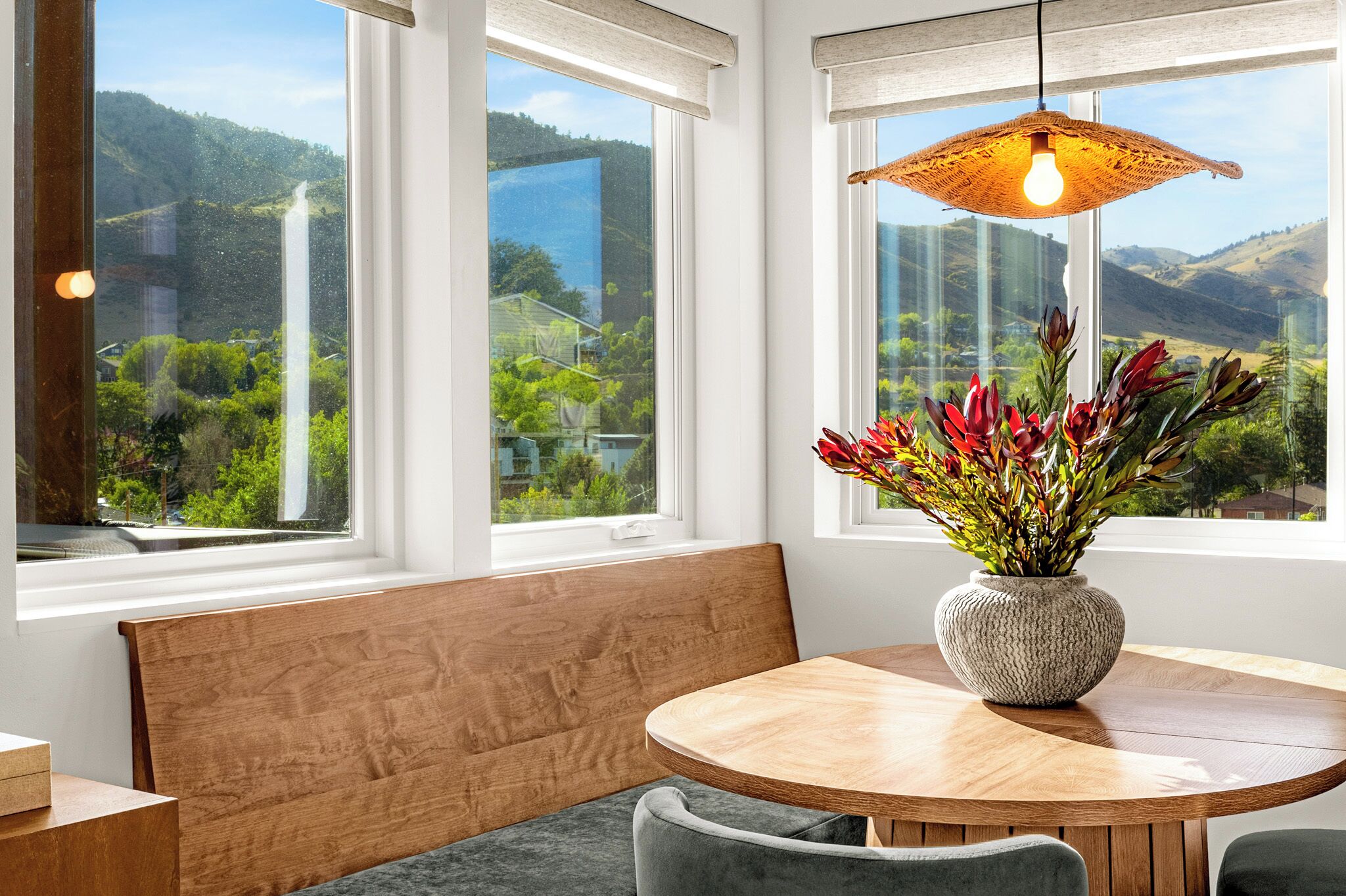 Une pièce avec une table ronde en bois, un banc et un vase rempli de fleurs. Il y a trois fenêtres qui donnent sur un paysage verdoyant et des collines.
