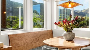 A room with a round wooden table, a bench, and a vase with flowers. There are three windows with views of greenery and hills. - Hot Tub, Mt Views, Luxury New build in Golden (Golden)